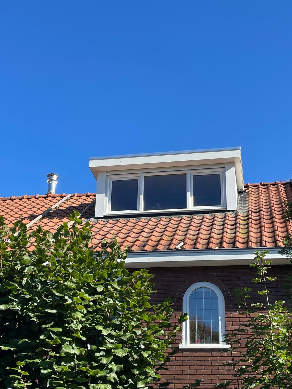 Witte dakkapel met drie ramen op een rood betegeld schuin dak, blauwe lucht en groen loof op de voorgrond.
