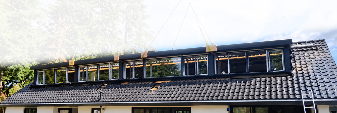Langwerpige dakkapel met meerdere ramen wordt gemonteerd op een schuin dak van een woning met zwarte dakpannen.